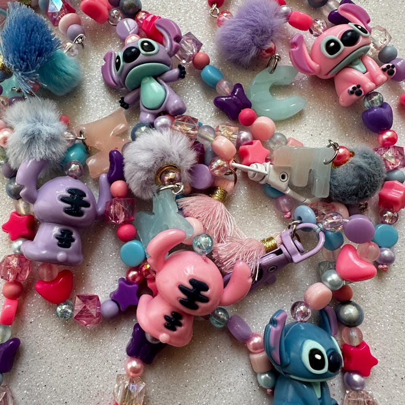 May include: A collection of colorful beaded accessories featuring cartoon character figures. The accessories include pink, purple, and blue Stitch figurines, star-shaped beads, and fluffy pom-poms. The beads are in various shapes and sizes, with a mix of pastel and vibrant colors.
