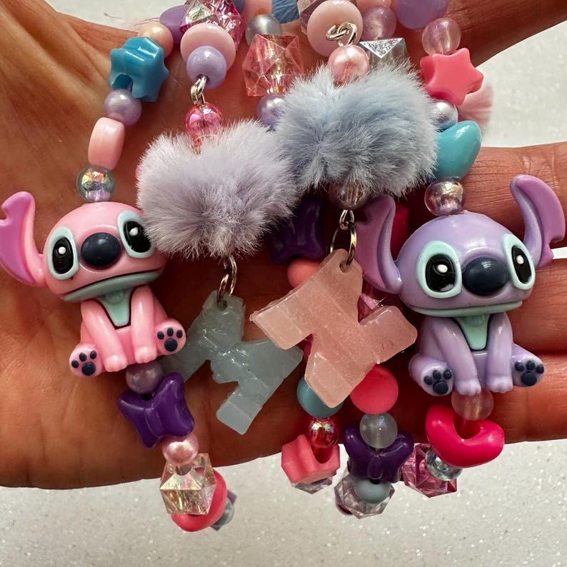 May include: Close-up of beaded accessories featuring cartoon character Stitch charms in pink and purple. The accessories include various beads in pastel colors, fluffy accents, and star-shaped charms. The overall theme is playful and whimsical.