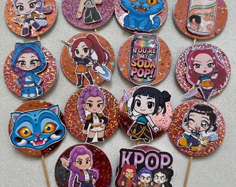 May include: A collection of decorative cupcake toppers featuring cartoon characters, a blue cat, and soda can designs. The toppers are round with glitter accents and attached to wooden sticks. Some toppers have the text "YOU'RE MY SODA POP!" and "KPOP DEMON HUNTERS".