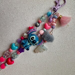 May include: A beaded keychain featuring a blue Stitch character charm, pink and red heart beads, and a pink tassel. The keychain also includes a gray pom-pom and various other colorful beads, with a purple clasp.