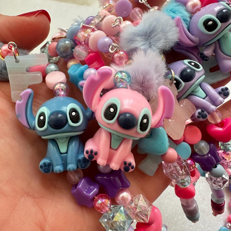 May include: A colorful beaded bracelet featuring Stitch and Angel figurines in blue and pink. The bracelet includes various beads in shades of pink, purple, blue, and clear, with star and heart-shaped accents and fluffy pom-poms.