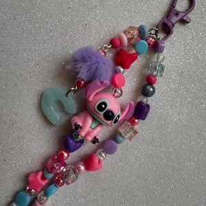 May include: A beaded keychain featuring a pink Stitch figurine, a light blue horseshoe charm, and a purple pom-pom. The keychain includes various beads in shades of pink, purple, blue, and clear, with a purple clasp.