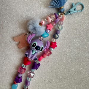 May include: A beaded keychain featuring a purple Stitch figurine, pink stars, and various colorful beads. The keychain includes a light blue tassel, a fluffy gray pom-pom, and a light blue clasp.