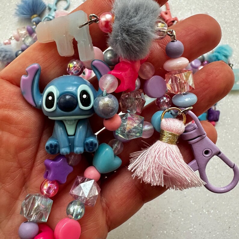 May include: A collection of beaded keychains in various colors. The keychains feature a blue Stitch figurine, a gray pom-pom, and a pink tassel. Beads are in shades of pink, purple, and clear.