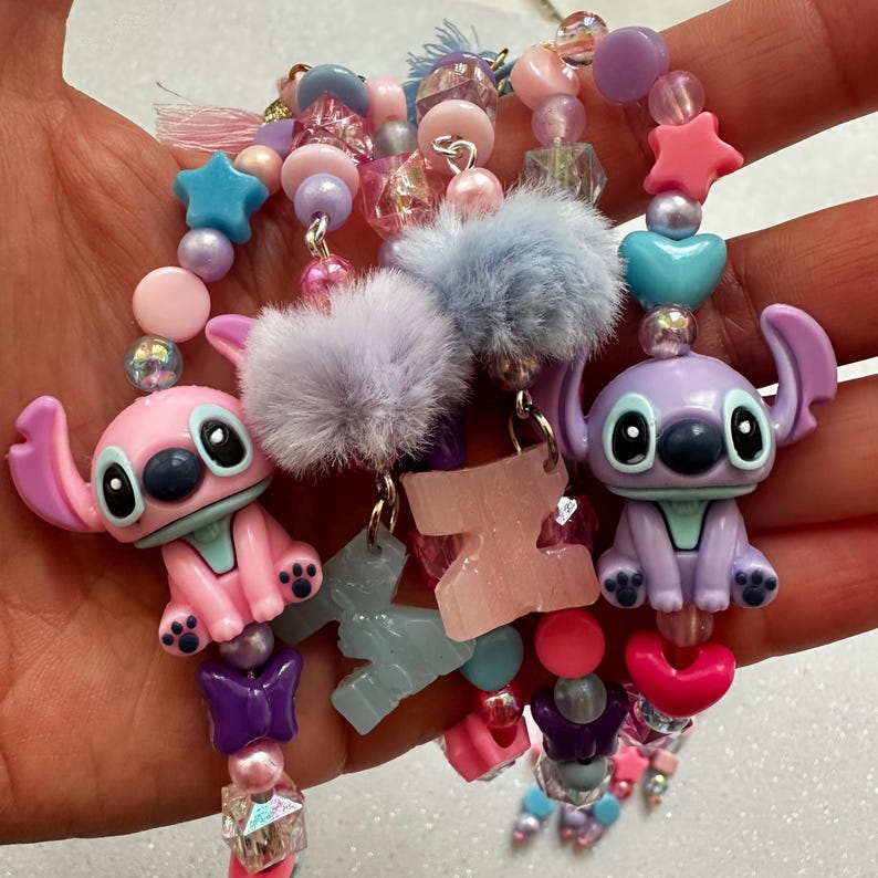 May include: A collection of beaded charms featuring pink and purple cartoon character figures, star, heart, and letter charms, and fluffy pom-poms. The beads are in shades of pink, purple, blue, and clear, creating a whimsical and colorful design.