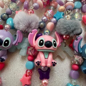 May include: Close-up of colorful beaded necklaces featuring cartoon character figures. The necklaces include pink, purple, blue, and clear beads, star shapes, and fluffy gray pom-poms. The cartoon characters are pink, purple, and blue.