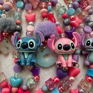 May include: A close-up of colorful beaded accessories featuring cartoon character figures. The beads are in shades of pink, blue, purple, and clear, with heart, star, and geometric shapes. The accessories include fuzzy pom-poms.