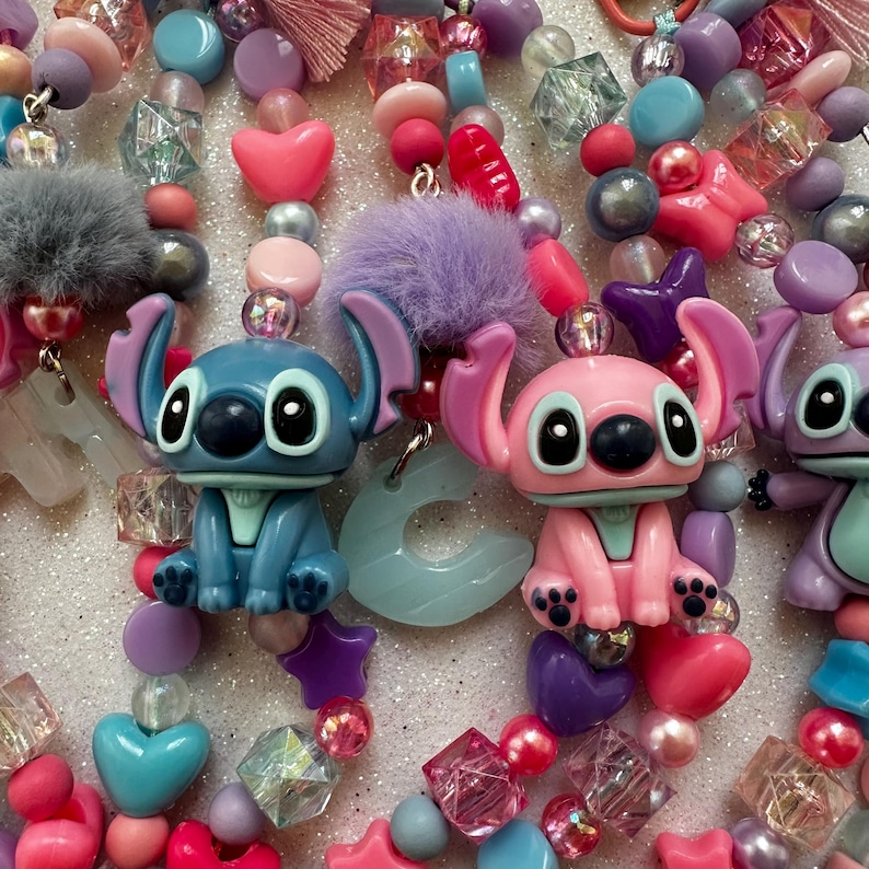 May include: A close-up of colorful beaded accessories featuring cartoon character figures. The beads are in shades of pink, blue, purple, and clear, with heart, star, and geometric shapes. The accessories include fuzzy pom-poms.