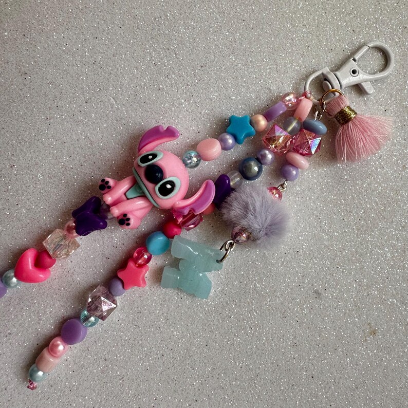 May include: A beaded keychain featuring a pink Stitch character, stars, hearts, and various beads in shades of pink, purple, and blue. Includes a light purple pom-pom and a pink tassel. The keychain has a silver clasp.