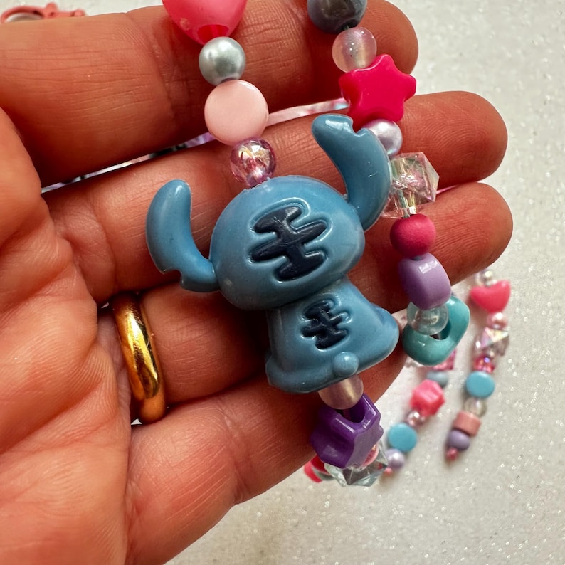 May include: A beaded bracelet featuring a light blue Stitch charm, pink star, heart, and round beads. The bracelet includes a variety of colorful beads in pink, purple, and clear. The bracelet is held in a hand.