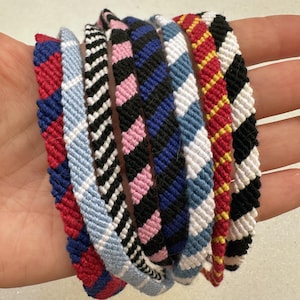 Peut inclure: Une collection de bracelets d'amitié colorés. Les bracelets présentent divers motifs, notamment des carreaux rouges et bleus, des chevrons noirs et blancs, des losanges roses et noirs et des carreaux bleus et blancs. Chaque bracelet est fait de fils tissés.