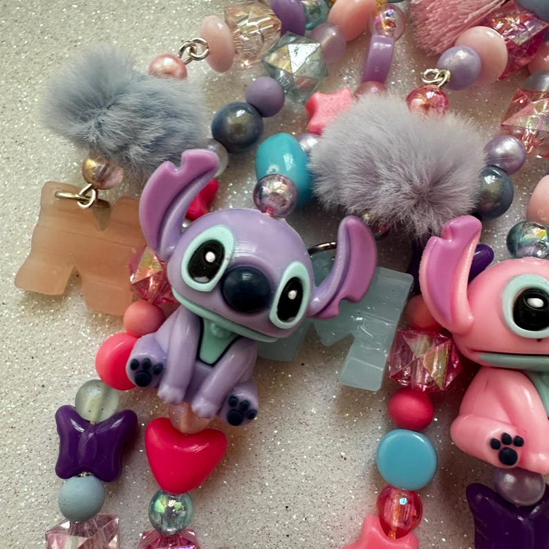 May include: Close-up of colorful beaded accessories featuring Stitch figurines, pom-poms, and various shaped beads. The color palette includes pink, purple, blue, and clear beads. The accessories are likely for a keychain or necklace.