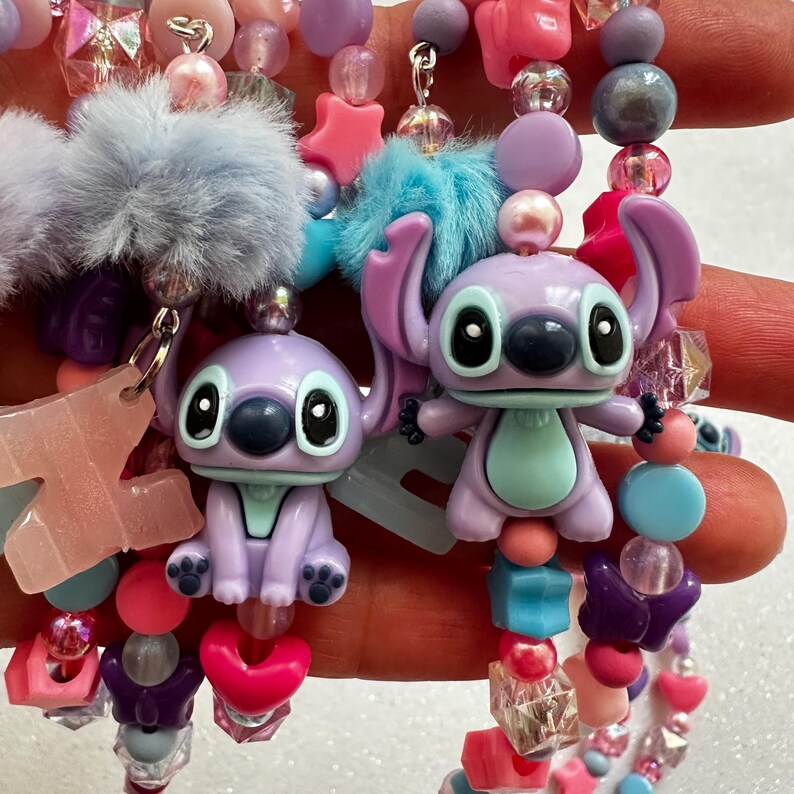 May include: A close-up of colorful beaded jewelry featuring Stitch character charms. The bracelets and necklaces include pink, purple, blue, and clear beads, along with star and heart-shaped accents and fluffy pom-poms.