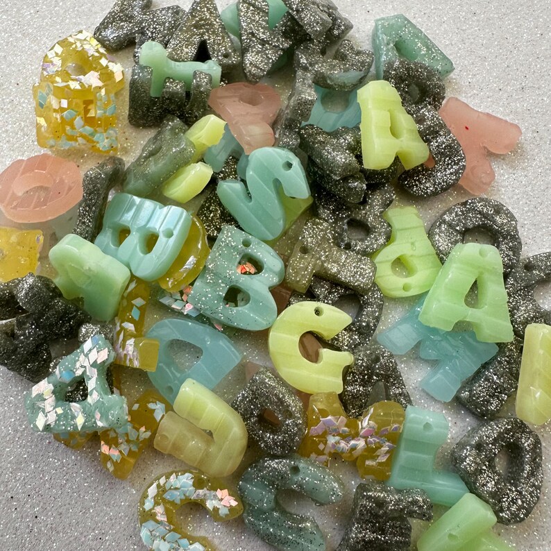May include: A collection of colorful resin alphabet letters in various shades of green, yellow, pink, and gray. Some letters are infused with glitter, creating a sparkling effect. The letters are scattered, showcasing different fonts and styles.