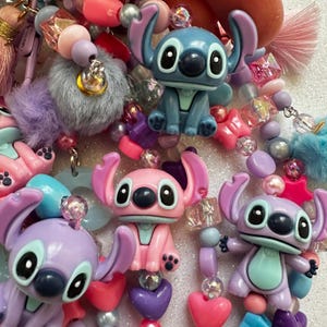 May include: A collection of colorful beaded accessories featuring Stitch figurines in blue, pink, and purple. The beads are in various shapes and colors, including hearts, stars, and round beads. The accessories also include fluffy pom-poms and tassels.