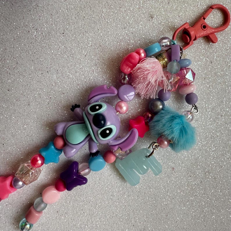 May include: A beaded keychain featuring a purple Stitch character charm, pink and blue star beads, and a light blue fluffy pom-pom. The keychain has a red clasp and various other colorful beads.