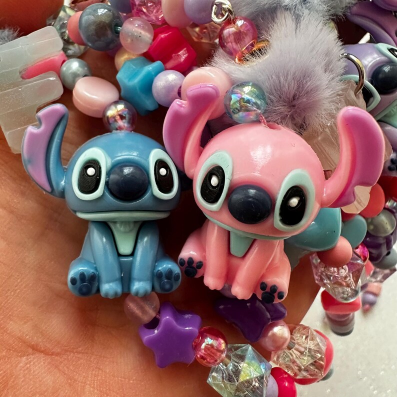 May include: Close-up of two cartoon character charms, one blue and one pink, with large black eyes and prominent ears. The charms are surrounded by colorful beads in various shapes and sizes, including stars and hearts. The overall theme is playful and whimsical.