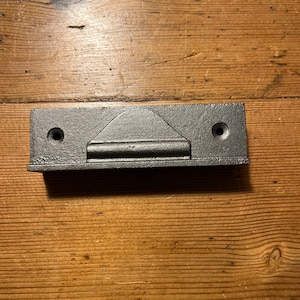 May include: Black metal door stop with a rounded top and two screw holes. The door stop is mounted on a wooden surface.