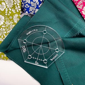 May include: A clear acrylic sewing tool with a circular design and measurements in inches, including "Height: 1.75 in" and "1 in". The tool is on a dark green fabric, with floral patterned fabric in the background. The tool is labeled "BOWE GOOSE".