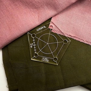 Könnte beinhalten: Ein klarer, fünfeckiger Quilt-Lineal mit einer 2,5 cm Messskala und dem Text "1/4 seam" und "Fowl Goose" aufgedruckt. Das Lineal liegt auf olivgrünem und altrosa Stoff.
