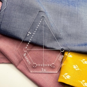 Può includere: Un righello per quilting in acrilico trasparente a forma esagonale, con misure in pollici. Il righello è contrassegnato con incrementi di 2 pollici (5,08 cm) e 1 pollice (2,54 cm), e un margine di cucitura di 3/8. Il righello è posto su tessuto blu, rosa e giallo.