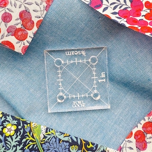 May include: A transparent, square quilting ruler, measuring 2.54 cm, placed on blue fabric. The ruler features markings for angles and seam allowances. The background includes floral patterned fabric in red, pink, and blue.