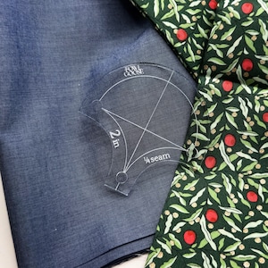 May include: A clear acrylic quilting template with the text "2 in" and "¼ seam" is placed on blue fabric. A patterned fabric with green leaves and red berries is visible in the background. The template is from "Fowl Goose".
