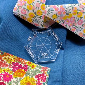 Op de afbeelding: Een doorzichtige zeshoekige quiltmal van acryl met afmetingen in cm, rustend op blauwe stof. Er zijn ook bloemmotiefstofstukken in roze, geel en oranje zichtbaar. De sjabloon is gemarkeerd met "1 in" en "¼ seam".