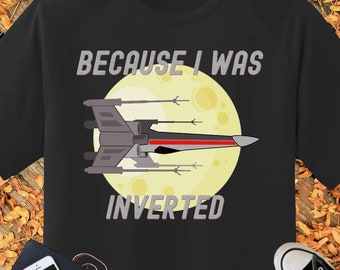 以下が含まれることがあります： 黒いTシャツ。黄色い月の前に宇宙船のグラフィックがあります。「BECAUSE I WAS INVERTED」の文字が宇宙船の上と下にグレーで印刷されています。シャツは、秋の葉のある木製の表面に表示されています。
