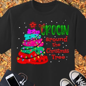 Könnte beinhalten: Schwarzes T-Shirt mit einem Weihnachtsbaum aus bunten Crocs-Schuhen. Der Text "Crocin around the Christmas Tree" ist auf dem Shirt aufgedruckt.