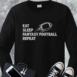 Puede incluir: Camiseta negra de manga larga con el texto plateado "EAT SLEEP FANTASY FOOTBALL REPEAT". Un gráfico de fútbol plateado está a la derecha del texto. Un smartphone, auriculares y zapatillas negras en primer plano.