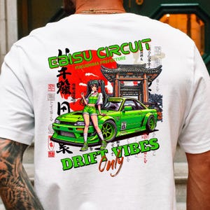 Könnte beinhalten: Weißes T-Shirt mit einem farbenfrohen Grafikdesign. Das Design zeigt den Text "EBISU CIRCUIT" und "DRIFT VIBES" in Grün, ein grünes Sportauto und einen Anime-Charakter. Japanischer Text und eine rote Sonne sind ebenfalls enthalten.