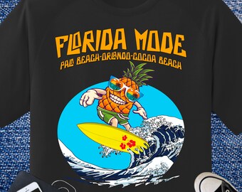 May include: Black t-shirt with a graphic of a surfing pineapple wearing sunglasses. The text "FLORIDA MODE" is above the pineapple, with "PALM BEACH-ORLANDO-COCOA BEACH" below it. The pineapple is riding a yellow surfboard on a blue wave.