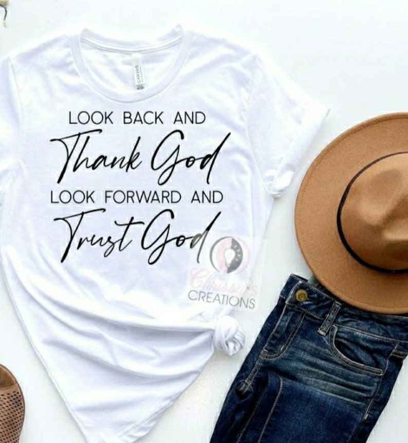 Pode incluir: Camiseta branca com o texto "Look back and thank God, look forward and trust God" em letras pretas.