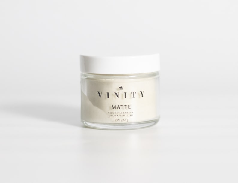Pode incluir: Um frasco de vidro transparente com tampa branca, contendo uma subst&acirc;ncia bege clara. O frasco tem um r&oacute;tulo preto com as palavras "VINITY" e "MATTE". O r&oacute;tulo tamb&eacute;m inclui o texto "MEDIUM HOLD & NO SHINE" e "VEGAN & CRUELTY FREE". O frasco cont&eacute;m 56 g.