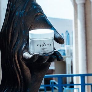 Pode incluir: Um frasco de vidro transparente com tampa branca, rotulado "VINITY MATTE", &eacute; segurado em uma m&atilde;o de cor bronze. O frasco cont&eacute;m uma subst&acirc;ncia de cor clara. O fundo apresenta um edif&iacute;cio com colunas brancas e uma balaustrada azul.