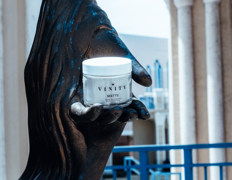 Pode incluir: Um frasco de vidro transparente com tampa branca, rotulado "VINITY MATTE", &eacute; segurado em uma m&atilde;o de cor bronze. O frasco cont&eacute;m uma subst&acirc;ncia de cor clara. O fundo apresenta um edif&iacute;cio com colunas brancas e uma balaustrada azul.