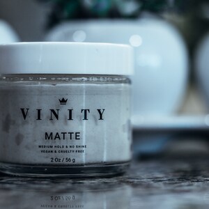 Pode incluir: Um frasco de vidro transparente com tampa branca, rotulado "VINITY MATTE". Cont&eacute;m uma subst&acirc;ncia clara e inclui "VEGAN & CRUELTY FREE", com um peso l&iacute;quido de 56 g. Vasos brancos desfocados no fundo.
