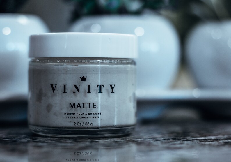 Pode incluir: Um frasco de vidro transparente com tampa branca, rotulado "VINITY MATTE". Cont&eacute;m uma subst&acirc;ncia clara e inclui "VEGAN & CRUELTY FREE", com um peso l&iacute;quido de 56 g. Vasos brancos desfocados no fundo.