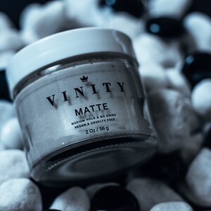 Pode incluir: Um frasco de produto de styling capilar Vinity Matte. O frasco de vidro transparente tem uma tampa branca e cont&eacute;m um creme de cor clara. O r&oacute;tulo diz "VINITY MATTE" com texto adicional indicando fixa&ccedil;&atilde;o m&eacute;dia, sem brilho e vegano e livre de crueldade. O frasco tem 56 g.