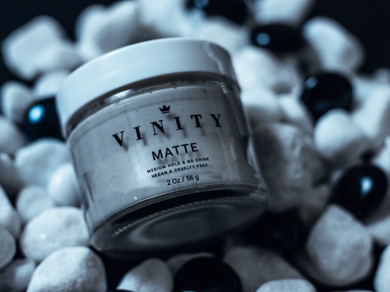 Pode incluir: Um frasco de produto de styling capilar Vinity Matte. O frasco de vidro transparente tem uma tampa branca e cont&eacute;m um creme de cor clara. O r&oacute;tulo diz "VINITY MATTE" com texto adicional indicando fixa&ccedil;&atilde;o m&eacute;dia, sem brilho e vegano e livre de crueldade. O frasco tem 56 g.