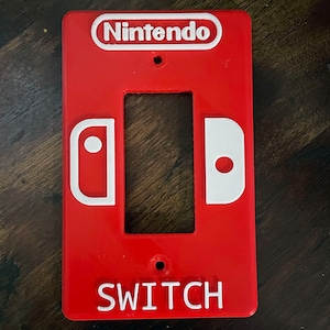 Peut inclure: Un cache-interrupteur en plastique rouge avec un logo Nintendo blanc et le mot "SWITCH" imprimé en dessous. Le cache comporte deux contours blancs des manettes Joy-Con de la Nintendo Switch.