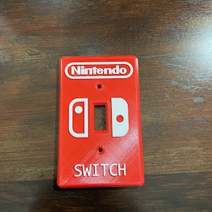 May include: Red Nintendo Switch light switch cover with white lettering and design. The word "Nintendo" is at the top, and the word "SWITCH" is at the bottom. The design includes the Switch controller icons.