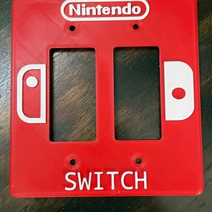 以下が含まれることがあります： 白い文字で「Nintendo Switch」と書かれた、2つの空白のスイッチプレートが付いた赤いプラスチック製の壁プレートです。