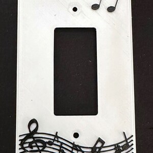May include: White plastic light switch cover with a black musical note design. The design features a treble clef and musical notes on a wavy line.