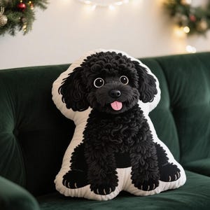 May include: A decorative pillow shaped like a black poodle with a white background. The pillow features a detailed design of the dog's fur, eyes, and tongue. The pillow is placed on a green velvet couch.