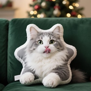 May include: A plush cat-shaped pillow with a gray and white color scheme, sitting on a green velvet couch. The cat pillow has a playful expression with its tongue sticking out. A Christmas tree is visible in the background.