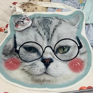 May include: A white tote bag with a blue background featuring a cartoon cat wearing round black glasses. The cat has gray and white fur and pink cheeks.