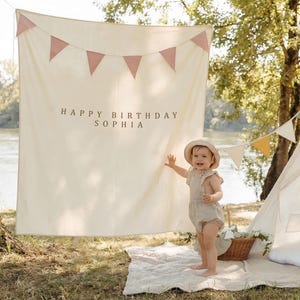 May include: A cream-colored banner with pink triangle flags and the text "HAPPY BIRTHDAY SOPHIA". A child in a beige outfit and hat stands in front of the banner. A picnic basket and tent are in the background.