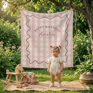 May include: A pink and white gingham backdrop with the text "Happy Birthday Amelia" hangs from a tree. A young child stands in front of the backdrop, wearing a light-colored romper. Wooden toys and a woven basket are on the ground.
